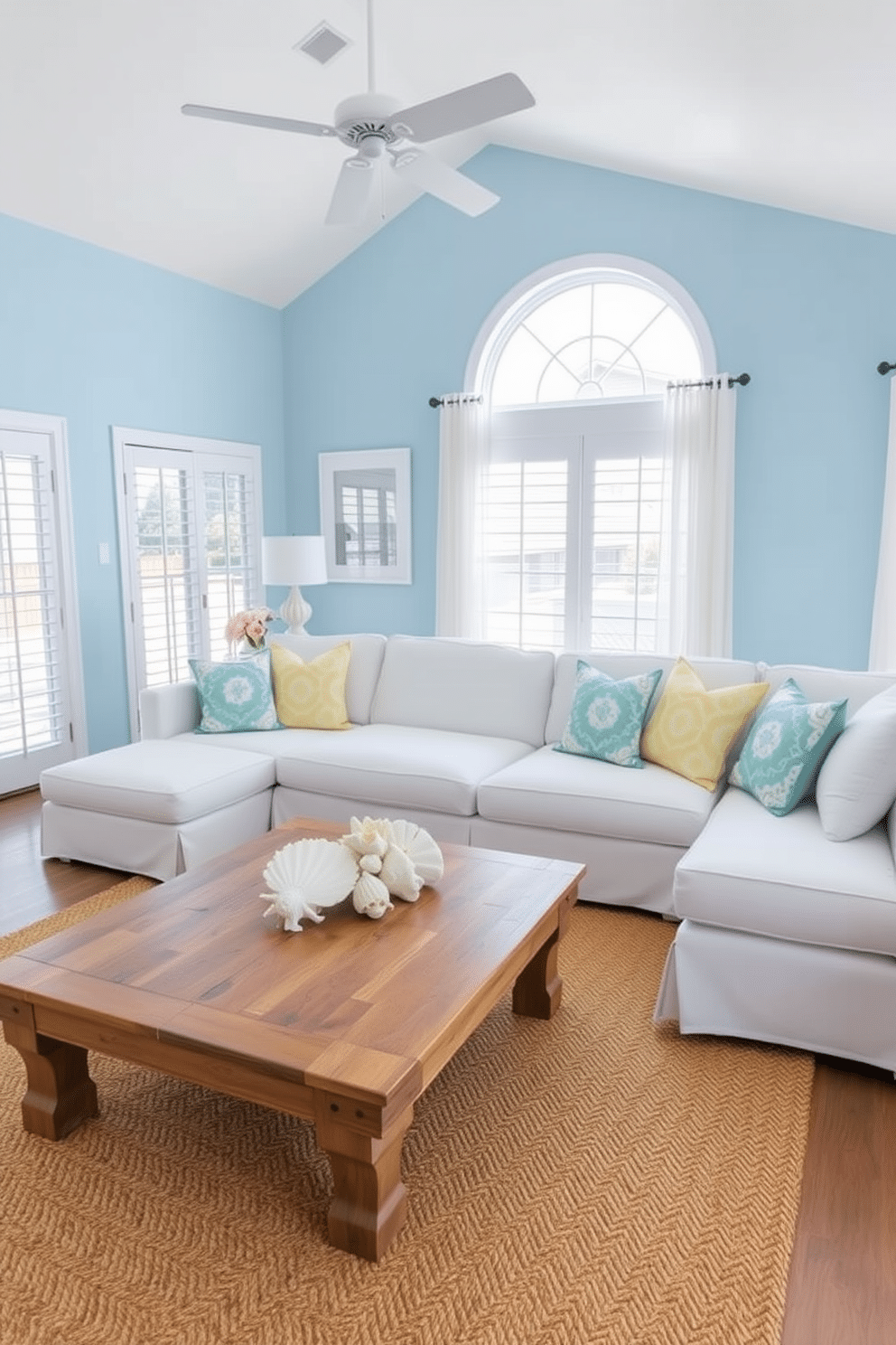 A serene living room with a beach-inspired color palette featuring soft blues and sandy beiges. The walls are painted in a light coastal blue, complemented by a large white sectional sofa adorned with vibrant throw pillows in ocean hues. Natural light floods the space through large windows dressed with sheer white curtains. A rustic wooden coffee table sits atop a woven jute rug, and seashell decor is artfully arranged on the table for a touch of summer charm.