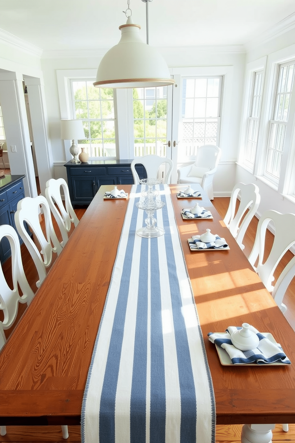 A bright summer dining room features a large wooden table adorned with nautical striped table runners and matching napkins. Surrounding the table are elegant white chairs that complement the coastal theme, and large windows allow natural light to flood the space.