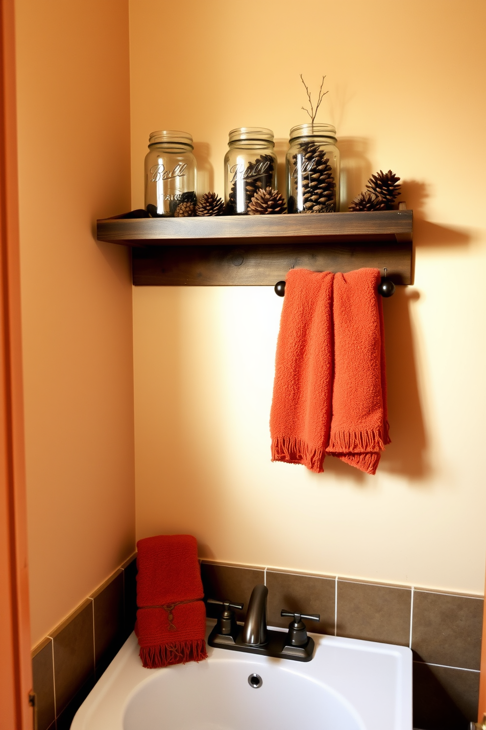 A cozy bathroom setting for Thanksgiving. Mason jars filled with pinecones are arranged on a wooden shelf above the sink, adding a rustic touch. The walls are painted in warm beige tones, complementing the autumn theme. Soft, fluffy towels in deep orange and brown hues hang neatly on a towel rack, enhancing the seasonal decor.