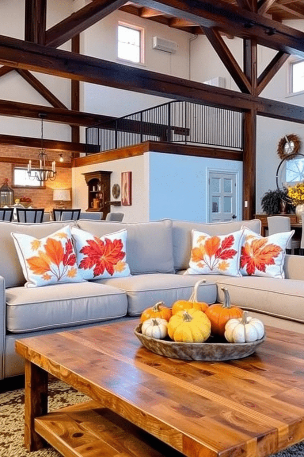 Throw pillows adorned with vibrant autumn motifs are scattered across a plush, neutral-colored sofa. The warm hues of orange, red, and gold create a cozy atmosphere, inviting guests to relax and enjoy the festive spirit. In the background, a beautifully decorated loft features rustic wooden beams and soft lighting, enhancing the seasonal charm. A centerpiece of pumpkins and gourds sits atop a reclaimed wood coffee table, completing the Thanksgiving aesthetic.