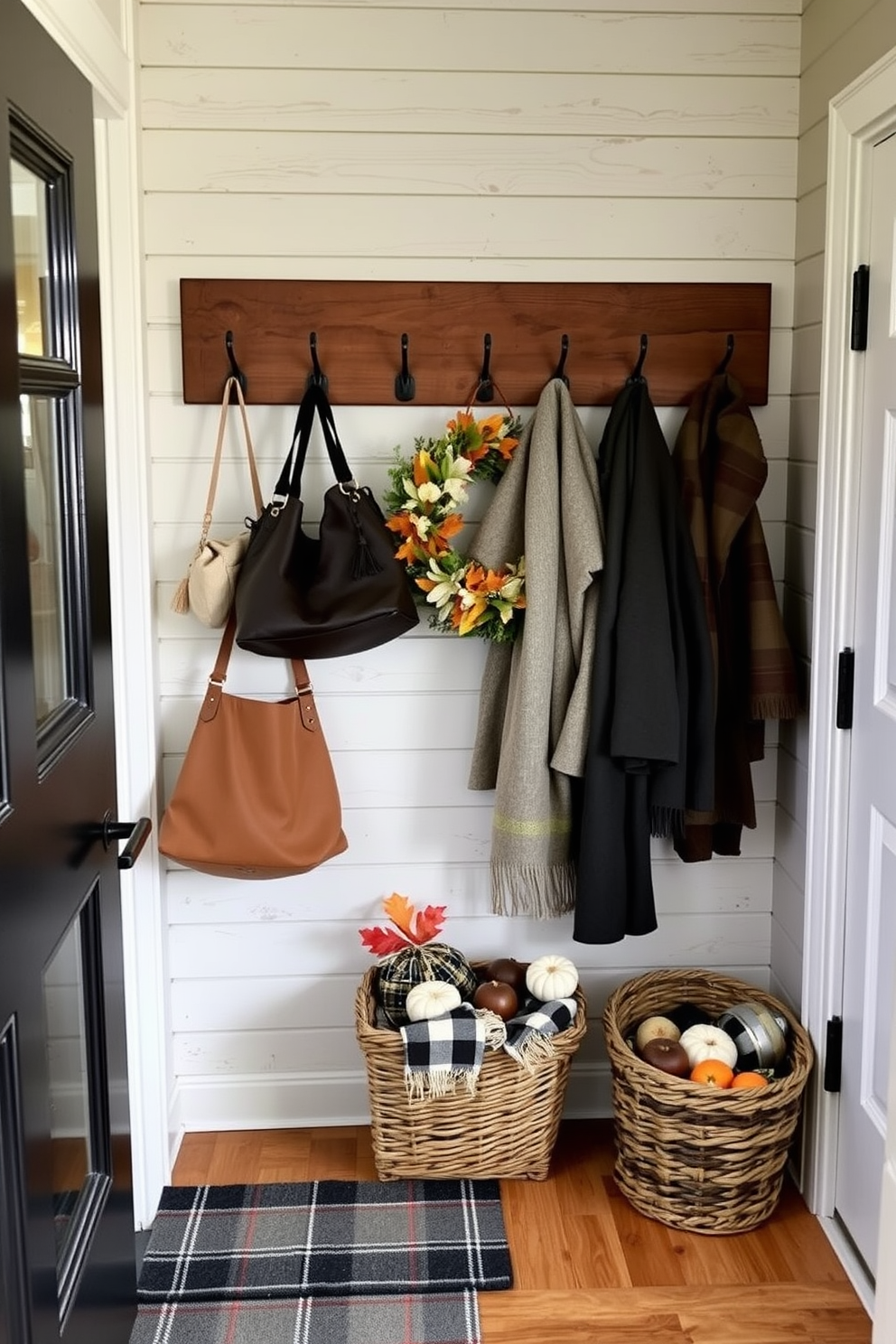 A cozy mudroom featuring rustic wooden hooks mounted on a weathered shiplap wall. The hooks are adorned with a mix of stylish bags and warm winter coats, creating an inviting and functional space. Decorative elements include a festive Thanksgiving wreath hanging on the door and a plaid runner on the floor. A basket filled with seasonal decor sits beside the entrance, adding warmth and charm to the mudroom.