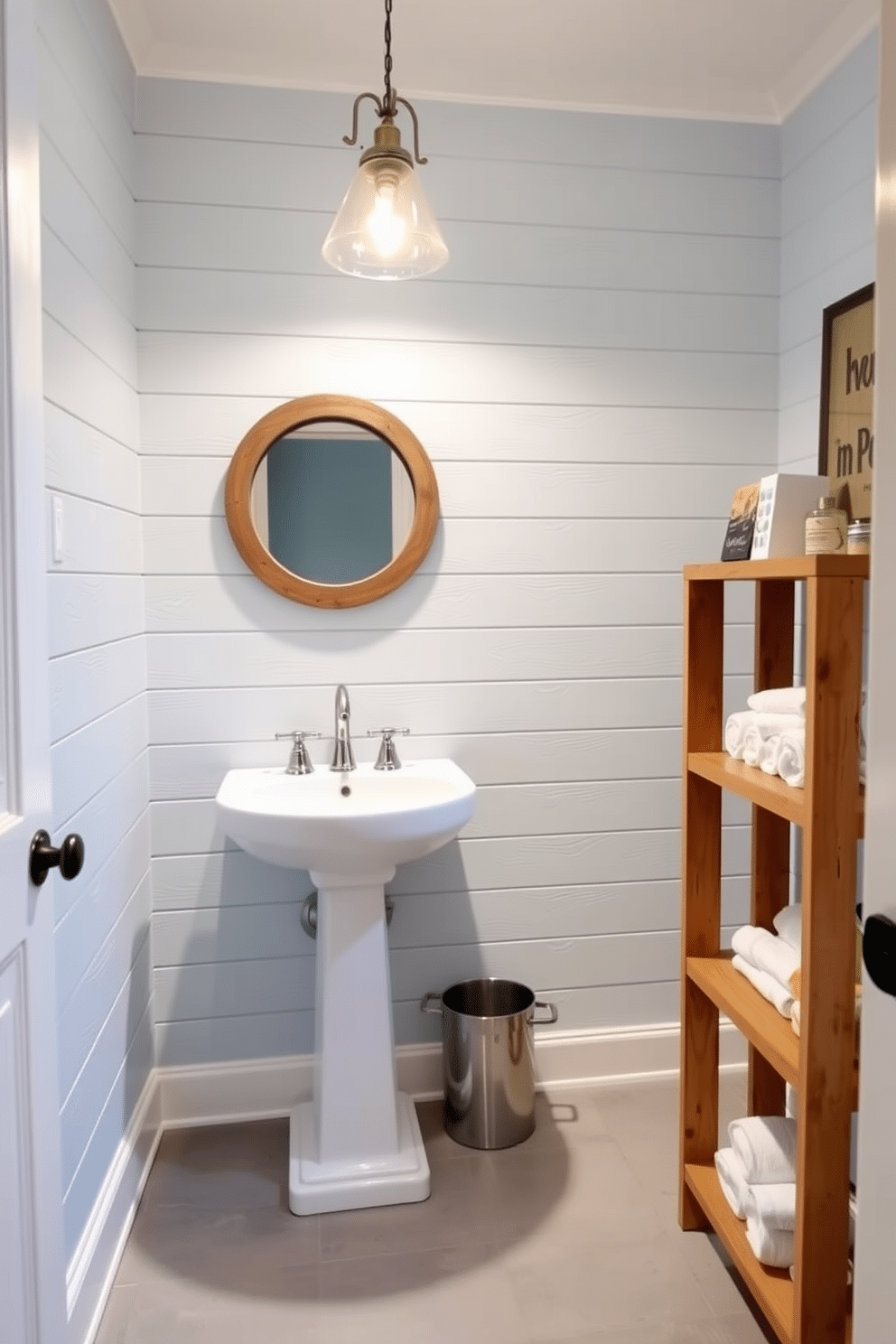 A stylish tiny powder room featuring an accent wall adorned with shiplap, painted in a soft, pale blue hue. The space includes a compact white pedestal sink and a round mirror with a rustic wooden frame, enhancing the room's charm. To the right, a small wooden shelf holds neatly rolled towels and decorative items. The floor is covered with light gray tiles, while a whimsical wall-mounted light fixture provides warm illumination, creating an inviting atmosphere.