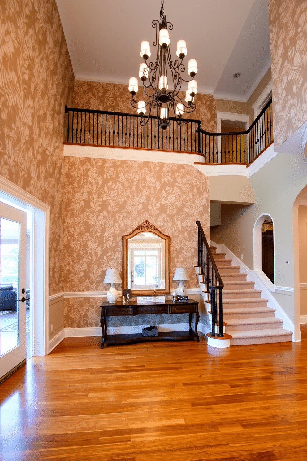 A stunning two-story foyer features an accent wall adorned with intricate floral wallpaper that adds depth and character to the space. The grand staircase, with its elegant wrought-iron railing, sweeps gracefully upwards, complemented by a beautiful chandelier that casts a warm glow throughout the area. The flooring consists of polished hardwood that contrasts beautifully with the soft, muted tones of the wallpaper. A large console table sits against the accent wall, adorned with decorative items and a striking mirror that enhances the foyer's spaciousness.