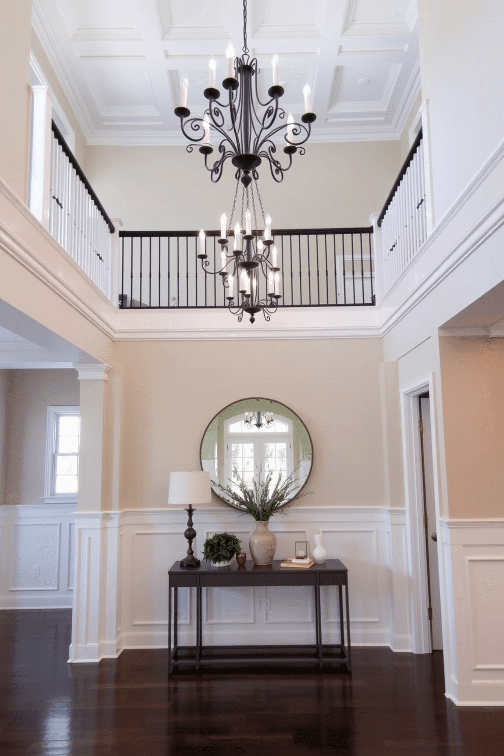 A stunning two-story foyer featuring a neutral palette that promotes a calming effect. The space is adorned with a grand chandelier hanging from a coffered ceiling, illuminating the elegant staircase with wrought iron balusters. The walls are painted in soft beige, complemented by white wainscoting that adds depth and sophistication. A large, round mirror is positioned above a sleek console table, which is decorated with a tasteful arrangement of greenery and decorative objects.