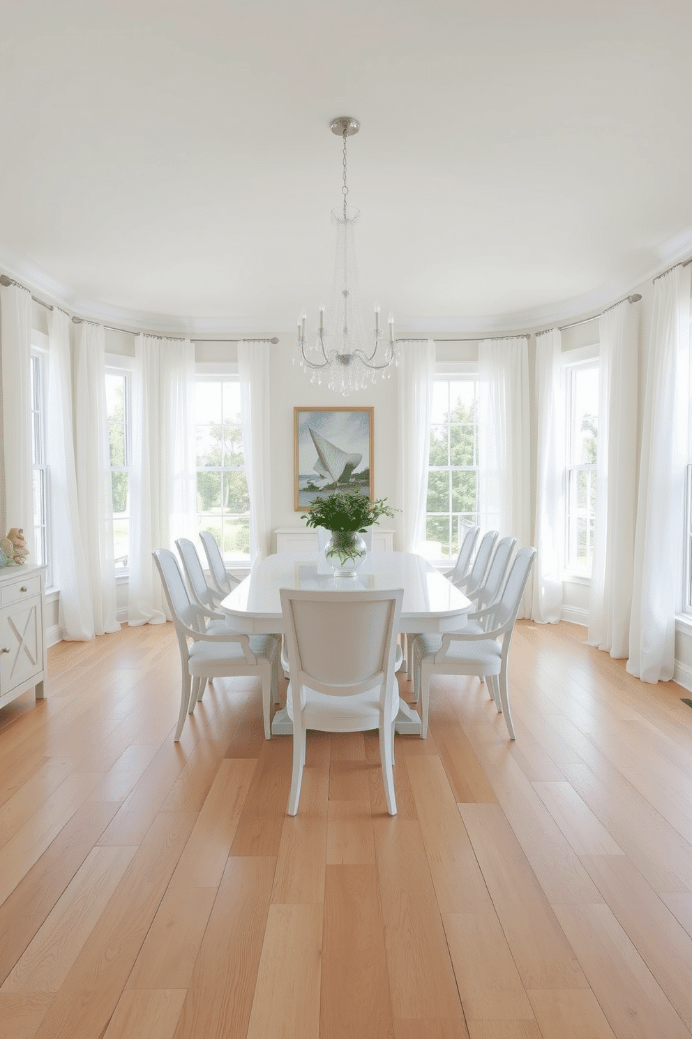 A bright and airy dining room featuring light wood floors that create a warm contrast with the crisp white decor. The room is adorned with a large white dining table surrounded by elegant white chairs, accented by a statement chandelier overhead. Large windows allow natural light to flood the space, enhancing the fresh and inviting atmosphere. A simple centerpiece of greenery adds a touch of nature, while soft white curtains frame the windows beautifully.