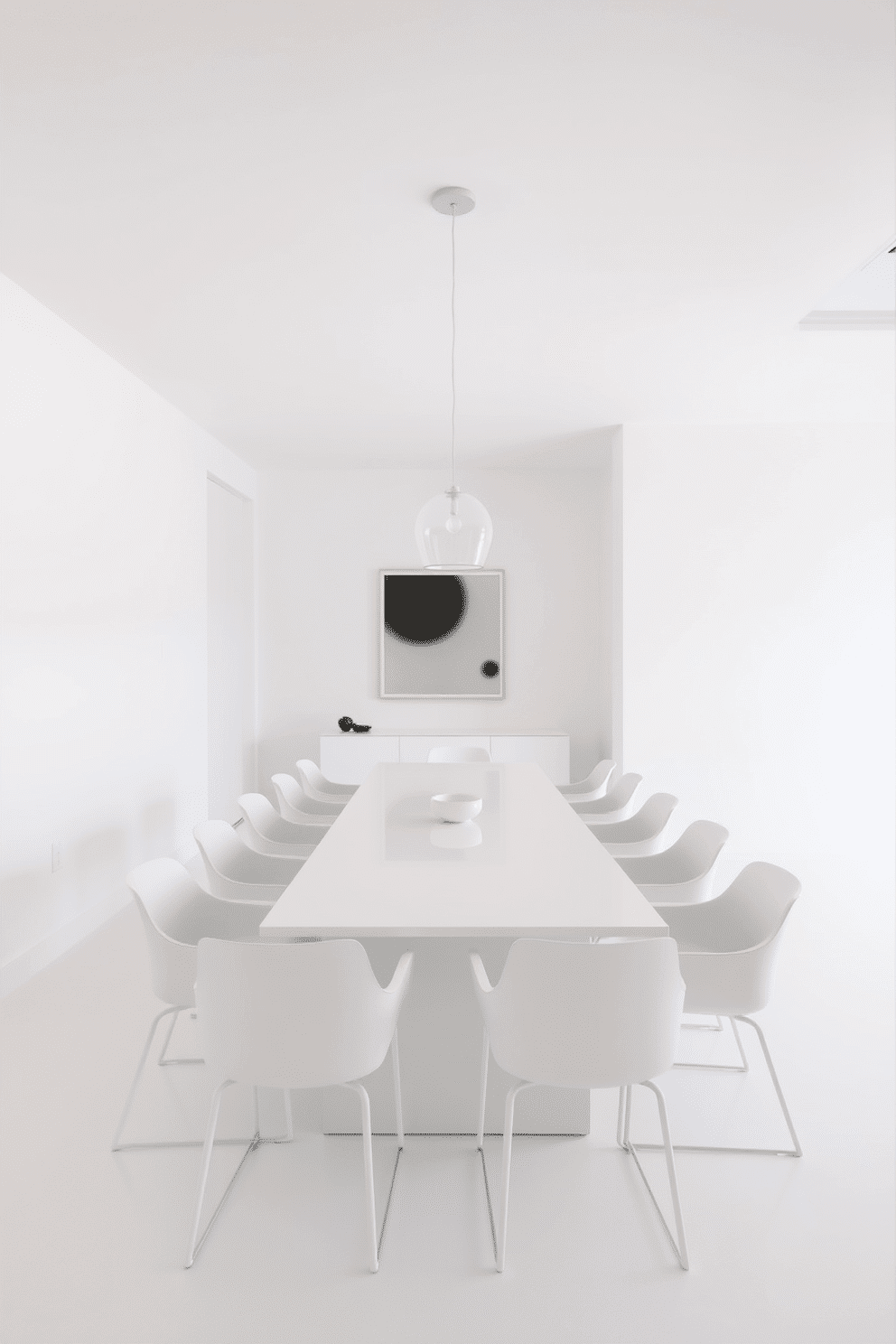 A minimalist dining room features sleek white furniture, including a long rectangular table surrounded by modern white chairs. The space is illuminated by a simple pendant light hanging above the table, creating an airy and open atmosphere. The walls are painted in a soft white hue, enhancing the room's brightness and making it feel more spacious. A single statement piece of artwork hangs on one wall, adding a touch of elegance without overwhelming the simplicity of the design.