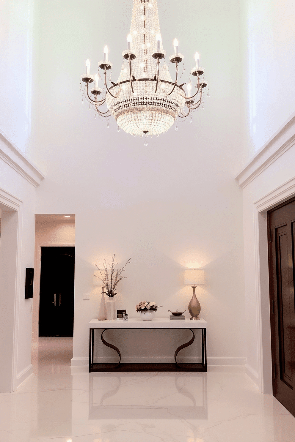 A grand entryway features an elegant crystal chandelier suspended from a high ceiling, casting a warm glow over the space. The white foyer is adorned with sleek marble flooring, complemented by a minimalist console table against the wall, topped with decorative accents and fresh flowers.