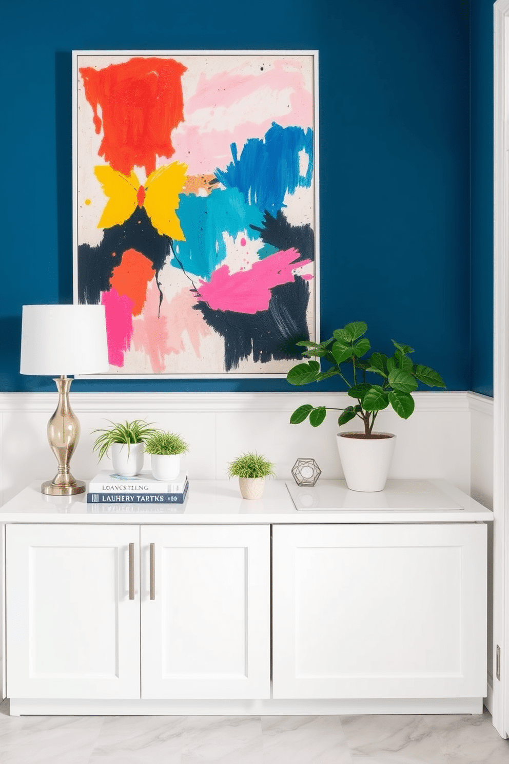 Bright artwork to liven the space. A large abstract painting with vibrant colors hangs prominently on the wall, infusing energy into the room. Below, a sleek, modern console table in white holds an arrangement of potted plants and decorative books, complementing the artwork beautifully. White laundry room design ideas. The space features crisp white cabinetry with brushed nickel hardware, providing ample storage while keeping the area looking clean and organized. A stylish folding station is integrated into the cabinetry, and the floor is adorned with light gray tiles for a fresh, airy feel.