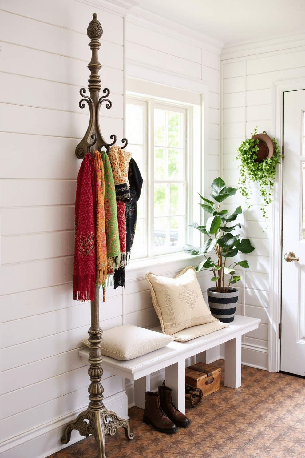 A vintage-style coat rack stands prominently against the wall, crafted from distressed wood with ornate detailing and a weathered finish. Below it, a collection of colorful scarves and hats hang, adding a personal touch to the space. The mudroom features a charming design with white shiplap walls and a durable, patterned tile floor that enhances the vintage aesthetic. A built-in bench with plush cushions sits beneath a window, providing a cozy spot to remove shoes, while potted plants add a touch of greenery to the inviting atmosphere.