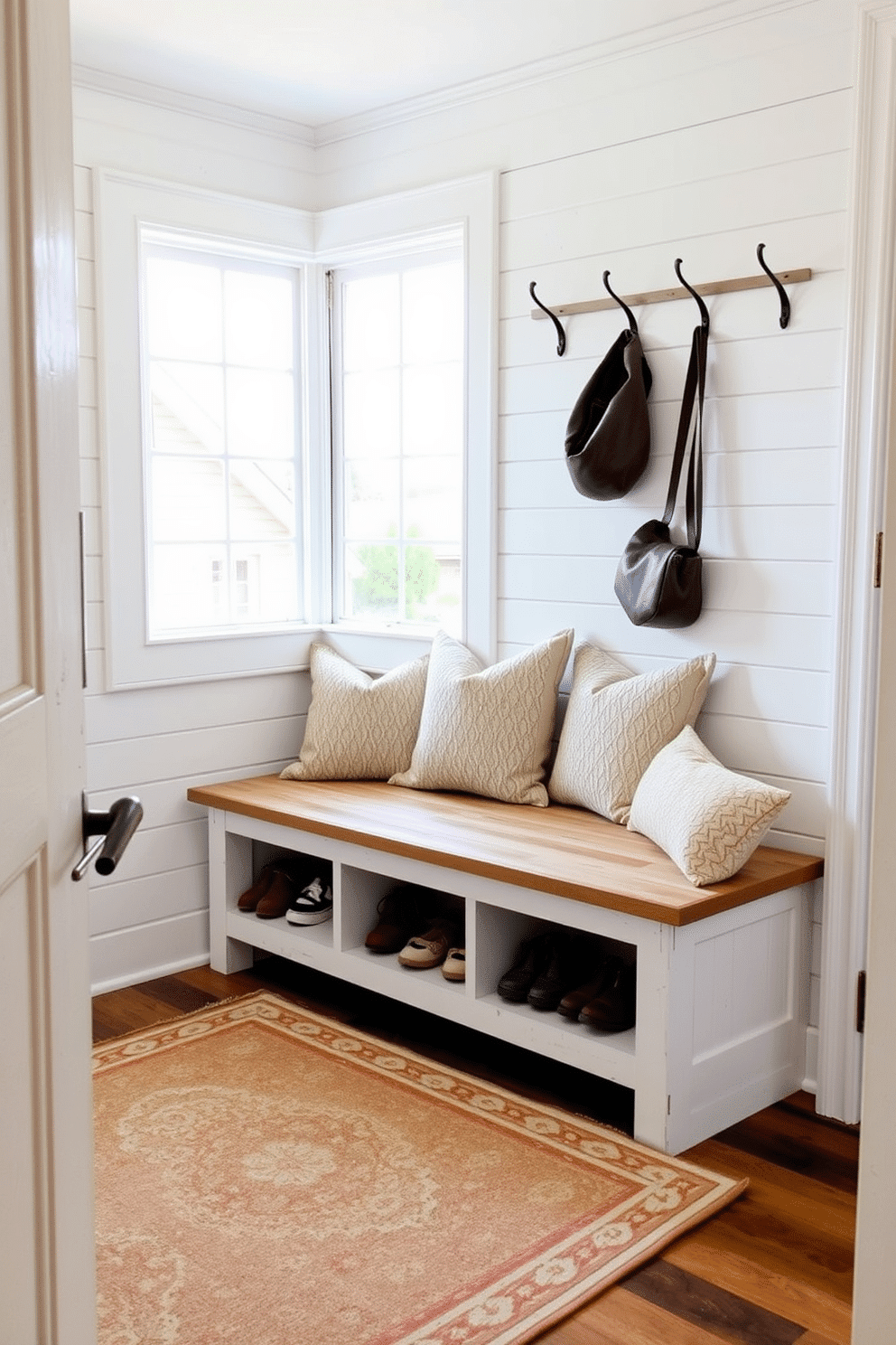 A charming farmhouse-style mudroom features a rustic wooden bench with built-in storage below, perfect for keeping shoes and outdoor gear organized. The space is adorned with shiplap walls painted in soft white, complemented by a vintage-inspired area rug that adds warmth and texture. Natural light floods the room through a large window, highlighting the bench's distressed finish and inviting cushions. Hooks line the wall above the bench, providing a practical yet stylish solution for hanging coats and bags.