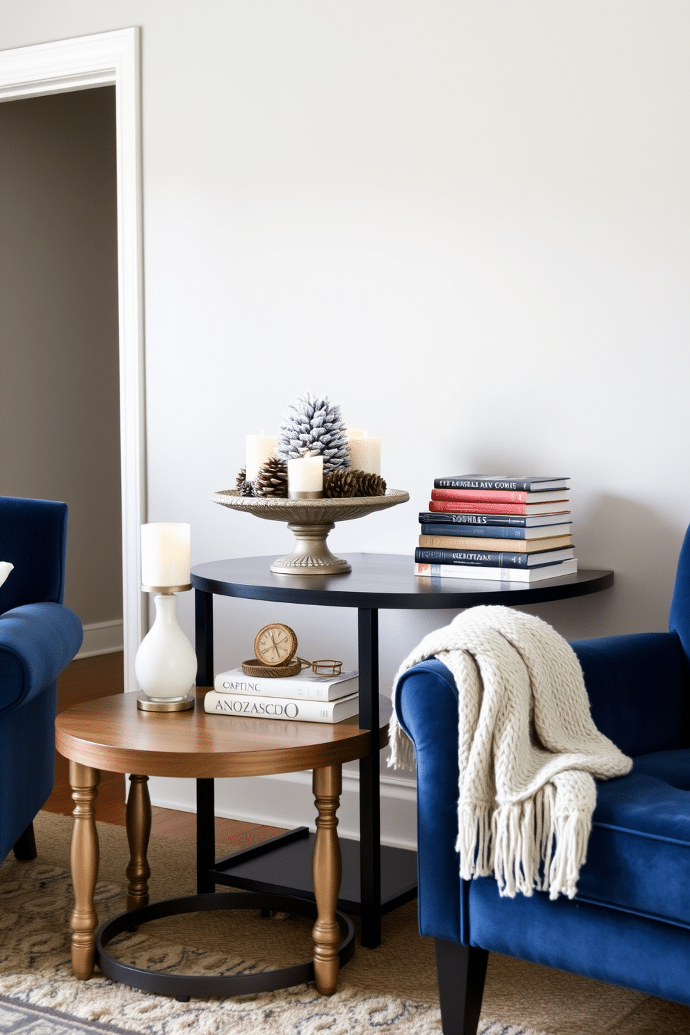 A cozy living room featuring accent tables adorned with winter decor pieces. One table is topped with a frosted pinecone centerpiece surrounded by small candles, while the other displays a stack of seasonal books and a knitted throw. The walls are painted in a soft gray, creating a warm backdrop for the decor. Plush armchairs in deep blue are positioned nearby, inviting relaxation during the chilly winter months.