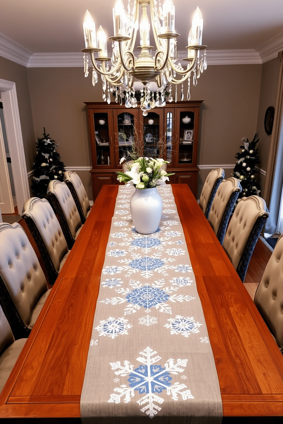 A winter-themed dining room featuring a long wooden table adorned with a beautiful table runner that showcases intricate snowflake patterns in shades of blue and white. Surrounding the table are elegant upholstered chairs with soft gray fabric, creating a cozy and inviting atmosphere for winter gatherings. The room is illuminated by a stunning chandelier with crystal accents that reflect the soft glow of candlelight. Frosted glass vases filled with evergreen branches and white flowers add a touch of natural beauty to the festive decor.