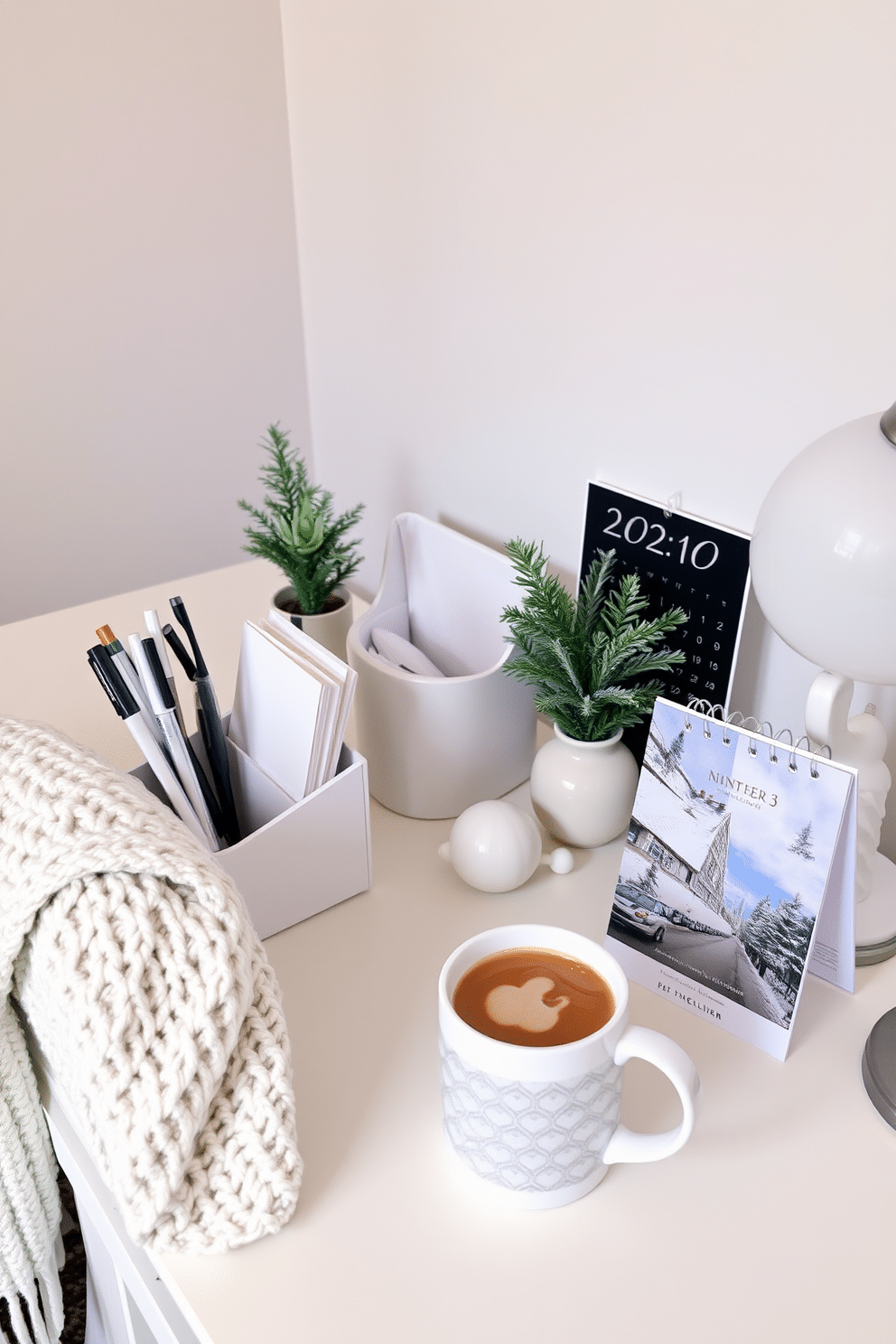 A winter-themed desk features elegant accessories in shades of white and silver. A sleek organizer holds pens and notepads, while a cozy knit blanket drapes over the chair. The desk is adorned with a small evergreen arrangement and a frosted glass lamp. A stylish calendar with winter imagery sits next to a steaming mug of cocoa.